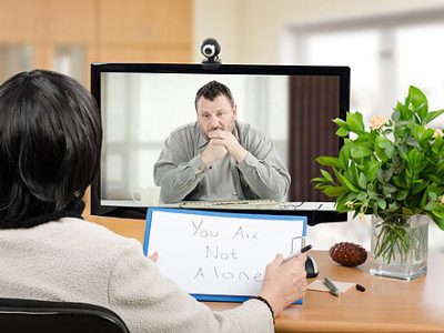 Middle aged man sitting in the monitor talks with psychotherapist via online video chat. He looking depressed. Black-haired psychiatrist holds written message for him - You are not alone. Horizontal shot indoors